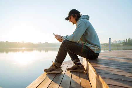 A hipster in a cap sits on a pier texting on his phone, a guy rests on the bank of the river, a man holding a smartphone, a city park. high quality photoの写真素材