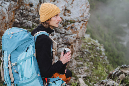 A climber in the mountains holds in his hand a flask with tea, a warm yellow hat, a turquoise...