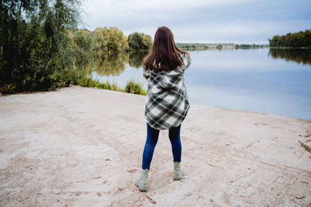 A girl in a pricked shirt and jeans is standing with her back on the pier, a woman looking into the distance at the water, a walk on the lake, a hipster on a walk. Casual style. High quality photoの写真素材