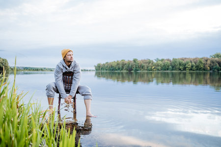 A man is sitting in the water on an old chair and looking at the lake, a guy is admiring the beautiful nature, a hipster is standing barefoot in the water. High quality photoの写真素材
