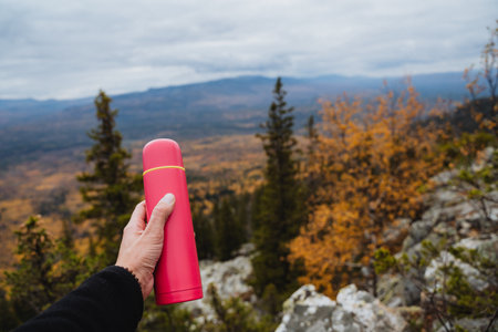 Pink flask in the hand of a man on the background of autumn forest, concept of drinking tea in the mountains in nature. Trekking in the mountains. High quality photoの写真素材