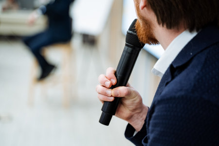A speaker holds a black microphone in his hand, a person makes a speech on stage, a lecture with a microphone, an object that transmits a voice. High quality photoの写真素材