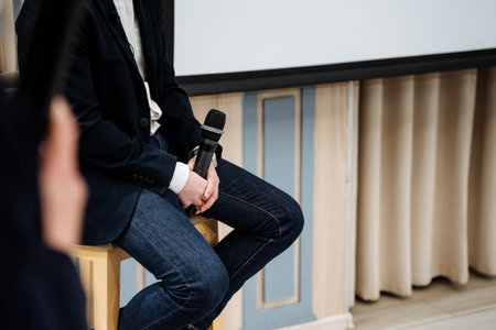 A man is sitting on a bar stool and holding a black microphone in his hands, a coach is giving a speech at a conference, a guy is giving a speech on stage. High quality photoの写真素材