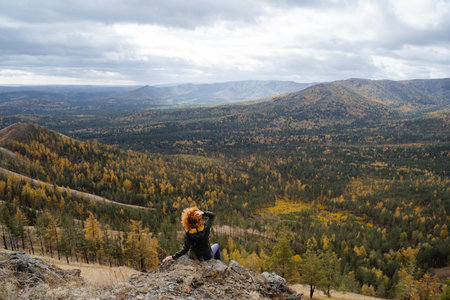 Girl with lush hairstyle sitting in the mountains and looking at the beautiful landscape, trekking in the mountains, autumn forest. High quality photoの写真素材