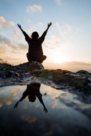 Reflection of the silhouette of a man in a puddle on a stone, back view of a girl sitting on a stone in the mountains, joyfully greeting the rising sun. High quality photoの写真素材