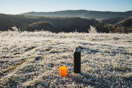 Black flask stands on the grass against the background of mountains, hot drink in thermal bottle, orange mug, drinking hot tea outdoors in winter. High quality photoの写真素材