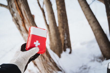 A hand in a glove is holding a red and white first aid kit in the winter forest. The kit is labeled with a white cross. The background is a blurred forest of trees with snow on the ground.の写真素材