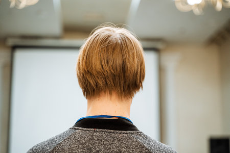 The person is standing with their back to the camera. They are wearing a gray shirt and have short blond hair. The background is a room with a projector screen. The person is alone in the room.の写真素材