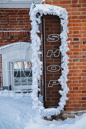 Snow-covered brick wall with a festive shop sign adorned with tinsel. The black metal sign reads shop in white letters, surrounded by snow on the ground. Winter decorations in the city.の写真素材