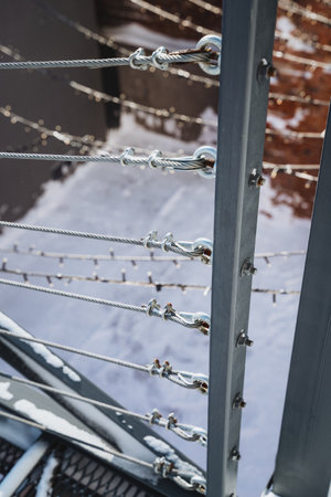 Close-up of gray metal railing with cable wires against snowy background, attached to post with...