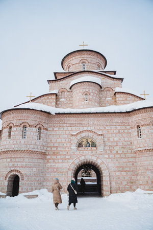 Two female tourists walking to old brick monastery in snowy landscape. Monastery made of red bricks,...