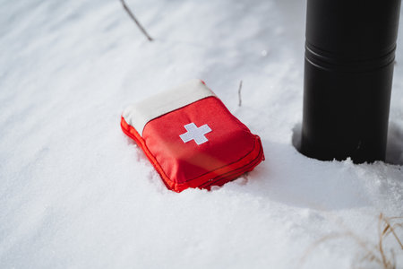 A small first aid kit with a red cross on it, laying in the snow near a black metal fence post. The kit is red and white, and it is about the size of a small wallet. It is covered in snow.の写真素材