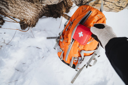 A colorful orange backpack with a red first aid kit hanging from a snowy tree. The first aid kit has a white cross. Open backpack with kit being removed in snowy forest.の写真素材