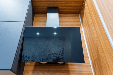 A rectangular black stove hood is suspended from the ceiling in a kitchen with hardwood flooring and...