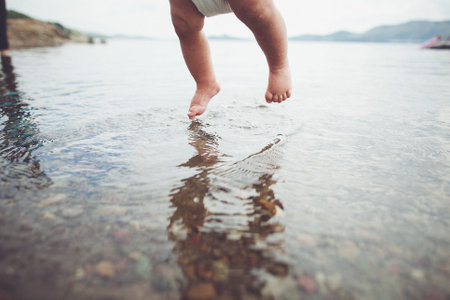 A young child wearing a diaper gleefully splashes into the refreshing liquid, showing a playful gesture at the edge of the beach or lake with their tiny human leg and footの写真素材