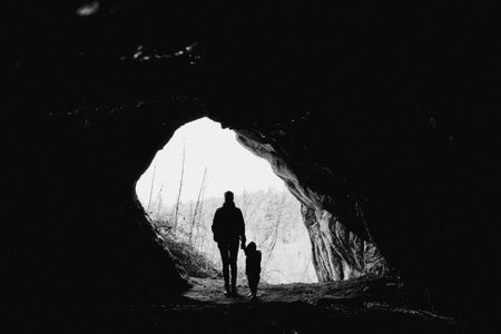 A man and a child are exploring a dark cave hand in hand, surrounded by coastal and oceanic landforms. The cave formations create a dramatic landscape with tints and shades of monochrome photographyの写真素材