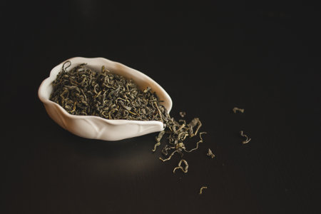 A white bowl filled with terrestrial plant green tea leaves resting on a black table, creating a...