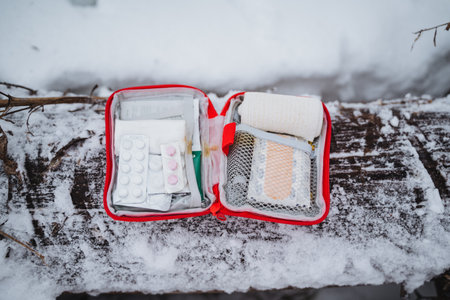 A first aid kit is placed on a snowcovered bench outside a building, with freezing temperatures. The sky is overcast, and nearby automotive mirrors and lighting are dusted with snowの写真素材