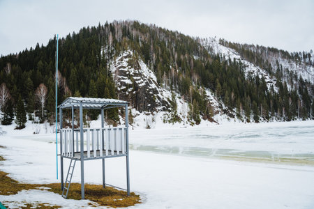 A lifeguard tower stands on the shore of a snowcovered lake, surrounded by mountainous landforms, trees, and a freezing natural landscapeの写真素材