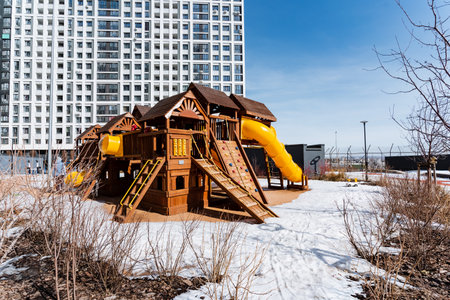 A wooden playground with a yellow slide covered in snow sits in front of a building. The white sky contrasts with the vibrant colors of the play structure, creating a picturesque sceneの写真素材