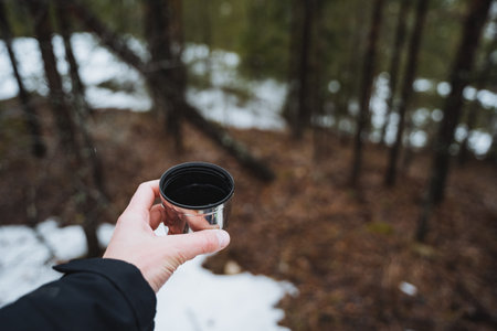 Cup of coffee in the hand of a man on the background of the forest, lying snow on the ground, drinking warming tea in the forest, metal cup on a hike. High quality photoの写真素材