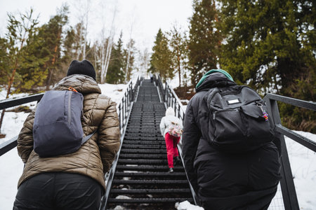 Two individuals are climbing a staircase covered in snow, surrounded by trees. They are bundled up in helmets and warm clothing, braving the freezing temperatures for a leisurely walkの写真素材