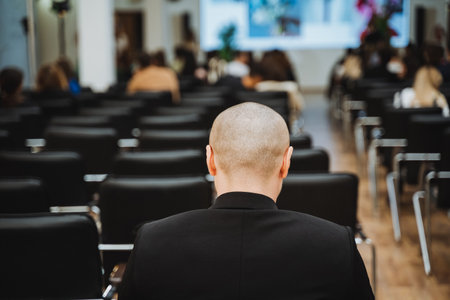 A bald man in formal wear is seated in a conference hall, attentively listening to a presentation during an official eventの写真素材