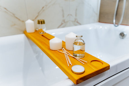 A wooden tray is placed on the edge of the bathtub in the bathroom, with liquid potential on its way to spill onto the floor due to its unstable position near the tapの写真素材
