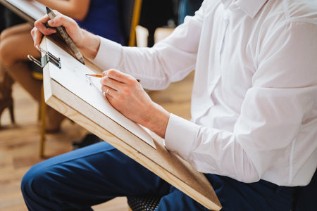A man is sitting at a hardwood desk, his thumb supporting his sleeve as he gestures with a pen on a piece of paper, focused on his drawing jobの写真素材