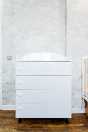 A rectangular white dresser on wheels made of hardwood is placed in the nursery next to a crib. The...