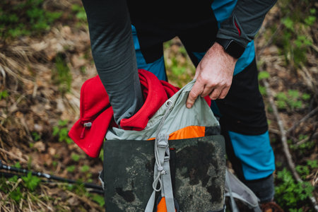 A person is leisurely packing a vibrant red jacket into their backpack amidst the lush green grass. The gesture suggests they are preparing for travel or recreation in natureの写真素材