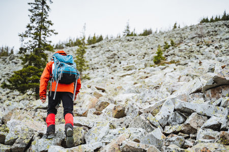 A hiker with a backpack is trekking up a rugged mountain slope surrounded by trees and a clear sky, embracing the beauty of the natural landscapeの写真素材