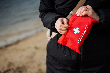 A woman with a red first aid kit stands on the beach, her hand on her waist, signaling readiness for...