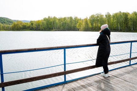 A woman enjoys the serene view of the lake from the wooden pier, with trees, water, and the open sky...