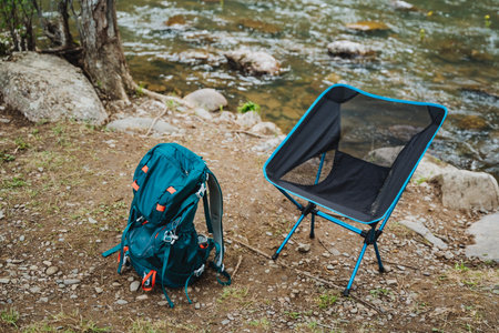 A chair and a backpack rest peacefully by the electric blue river, surrounded by lush green grass, tall trees, and beautiful woodland landscapeの写真素材