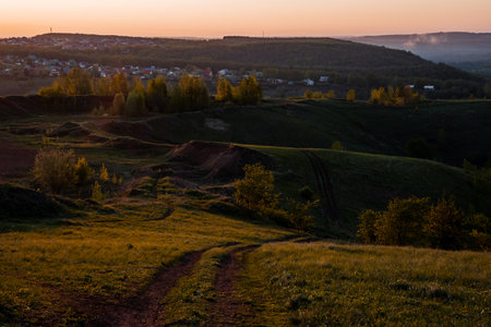 A grassy hillside under the colorful sunset sky, with a dirt road winding through the natural landscape of prairie grasses and distant horizonの写真素材