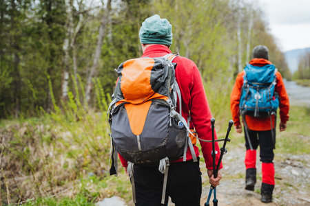 Two hikers carrying backpacks and hiking poles trek through a wooded trail, surrounded by trees and natural landscape, enjoying outdoor recreationの写真素材