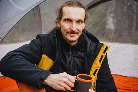 A happy man with a beard is sitting in a tent, wearing a jacket and personal protective equipment while holding a glass of coffee. He has a smile on his face and is ready to travelの写真素材