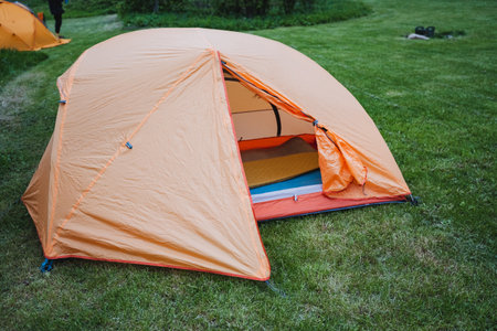 An inviting orange camping tent with its entrance unzipped, set up on a lush green lawn at a campsite. Ideal for outdoor adventures and nature enthusiasts seeking a comfortable home away from home.の写真素材