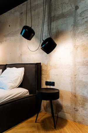Stylish modern bedroom showcasing a minimalist bedside table, with unique pendant lighting fixtures. The concrete wall adds a raw, industrial feel to the sleek, contemporary design.の写真素材