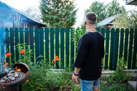 In a backyard with blooming flowers, a man is grilling food by a green fence. This tranquil outdoor setting is perfect for summer barbecues and relaxation moments for leisure and enjoymentの写真素材