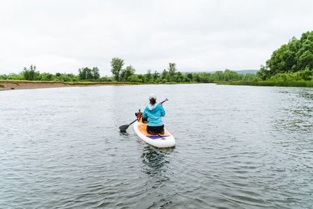 A kayaker in a blue jacket paddles on a peaceful river surrounded by green foliage, embracing the calmness of a cloudy day. Ideal for adventure, outdoor activities and nature scenes.の写真素材