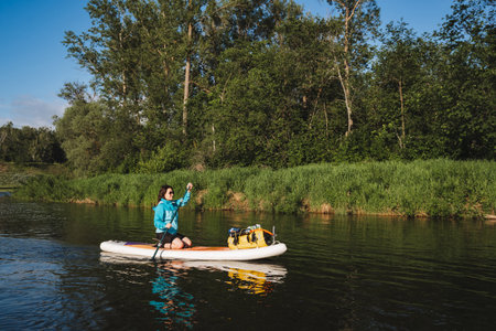 A woman and her dog paddleboarding on a serene river, surrounded by lush greenery. The peaceful...