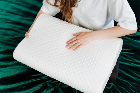 A woman gently holds a white memory foam pillow, showing its quilted texture, against a luxurious green velvet background. Perfect for advertisements about sleep and bedding products.の写真素材