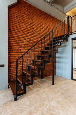 The chic black metal staircase in a modern industrial interior sets a striking contrast against the red brick wall, merging style and function to elevate the living area with sophisticationの写真素材