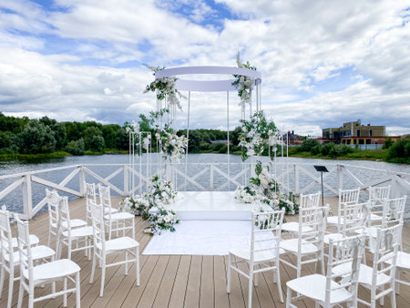 An enchanting outdoor wedding setup with a stunning floral arch, elegant white chairs by tranquil waters, and a picturesque sky, perfect for a memorable celebration of loveの写真素材