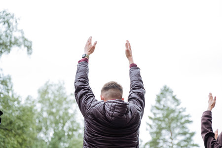 A group of joyful individuals raise their hands outdoors on grass under a tree, expressing joy and...