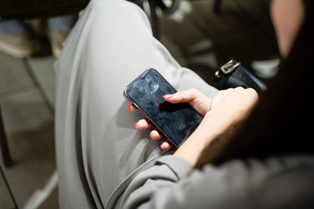 A woman sits comfortably in a cozy chair, casually holding her smartphone, deeply immersed in a...