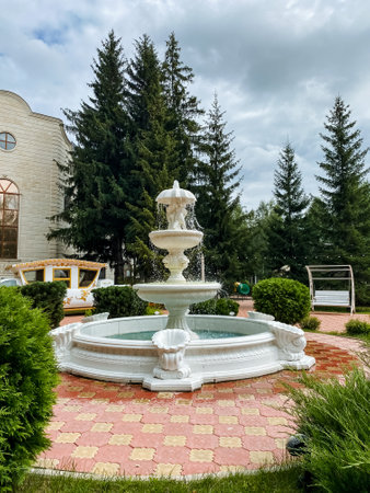 In the center of a beautifully designed park, there is an enchanting fountain which is elegantly situated and surrounded by a variety of lush trees that provide a serene atmosphereの写真素材