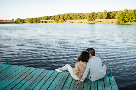 A loving couple comfortably relaxes on a beautiful wooden dock by the calm, serene waters, enjoying a stunning sunset and the incredibly peaceful atmosphere surrounding themの写真素材